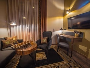 Een kamer met een zithoek, een ronde salontafel met schaakstukken, een bureau met een stoel en een aan de muur bevestigde televisie.