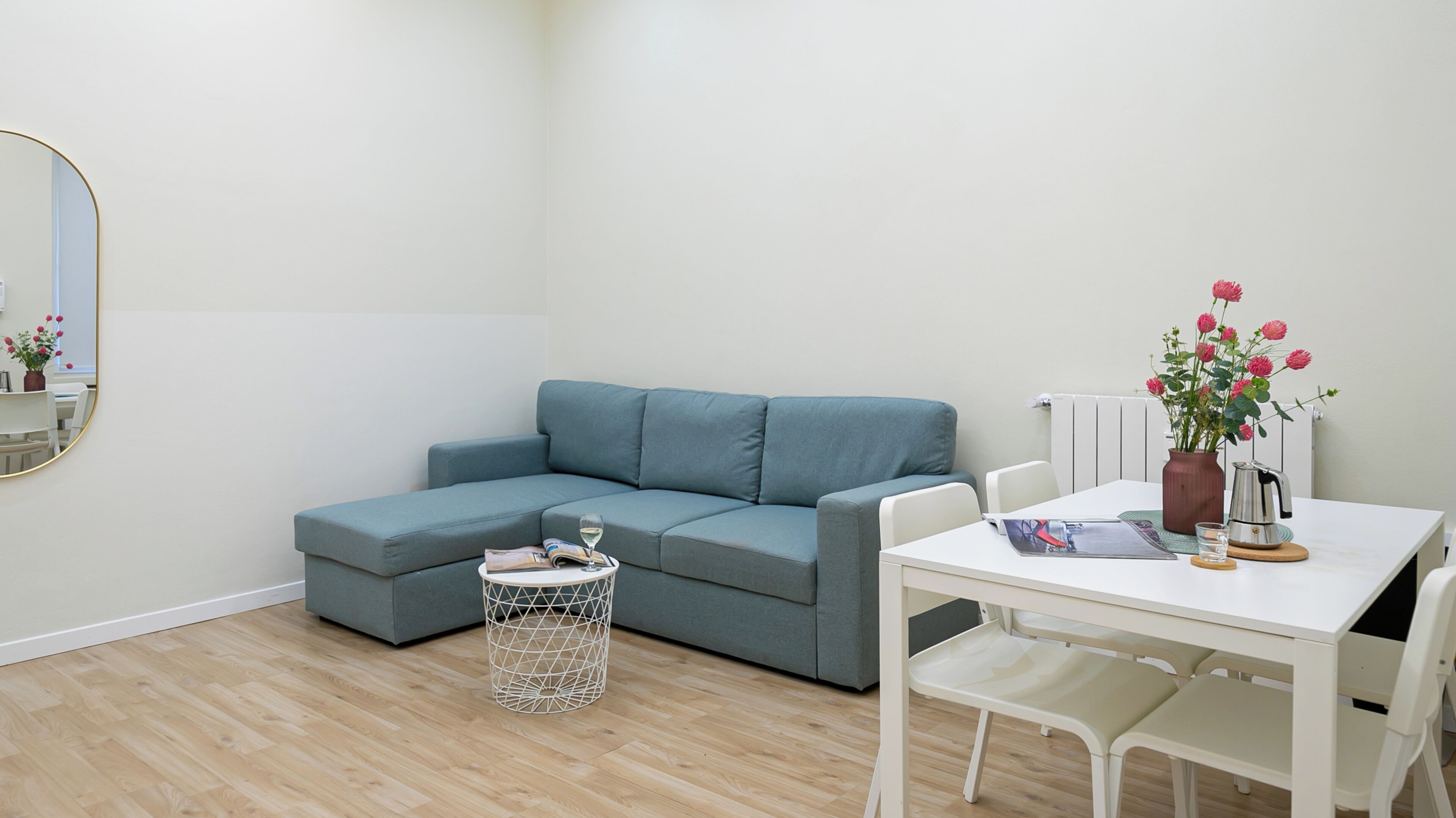 Un salon moderne avec un canapé d’angle gris, une petite table d’appoint ronde, une table à manger blanche avec des chaises et une plante en pot aux fleurs rouges.