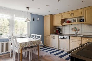 A kitchen with wooden cabinets, a dining table with chairs, a coffee maker, and a window with curtains. - 3BR In Kosta - Fire pit, gathering room, Lake &Pub (Källeskruv)