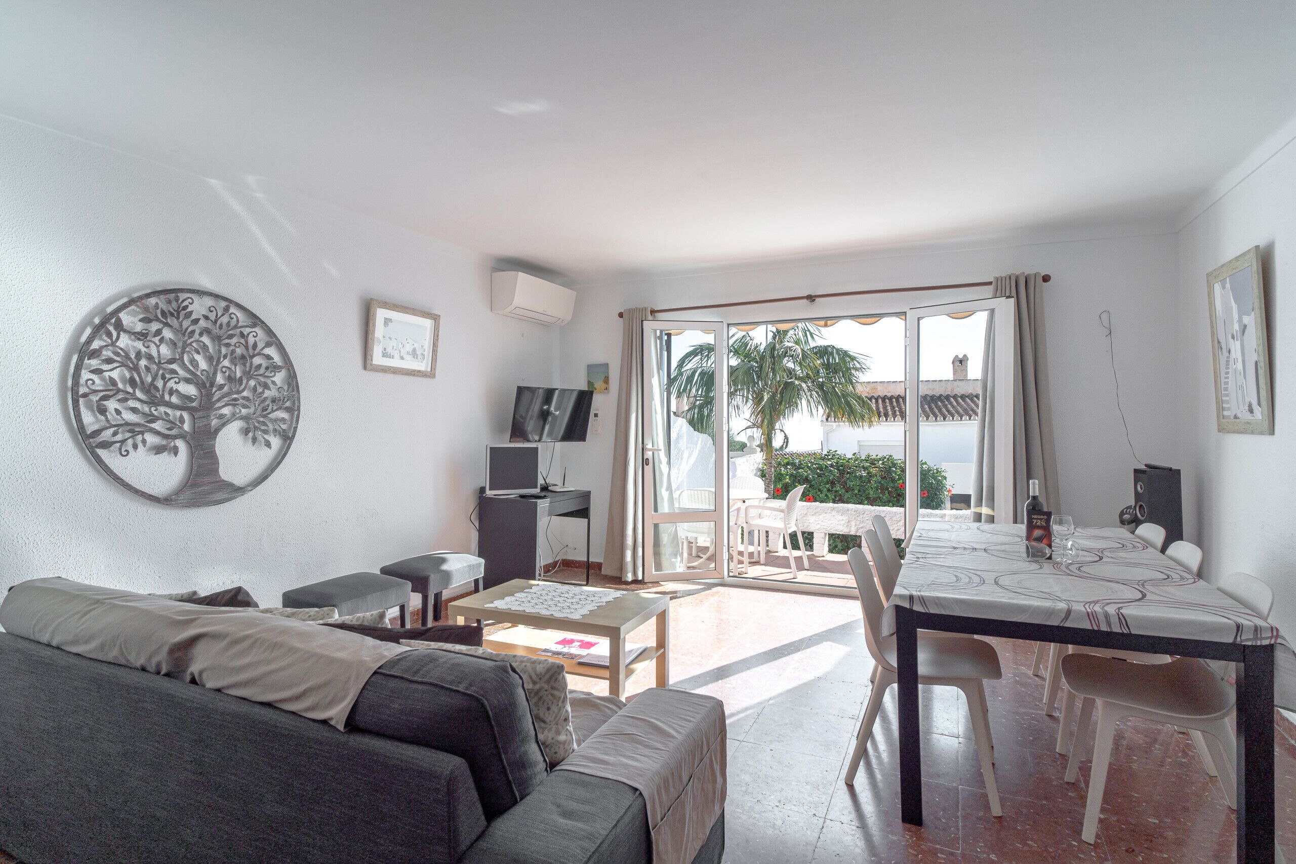 Een woonkamer met een bank, salontafel, televisie en een eettafel met stoelen. Er is een balkon met uitzicht op palmbomen en een gebouw.