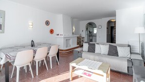 En stue med sofa, spisebord med stole, pejs og et køkken i baggrunden.