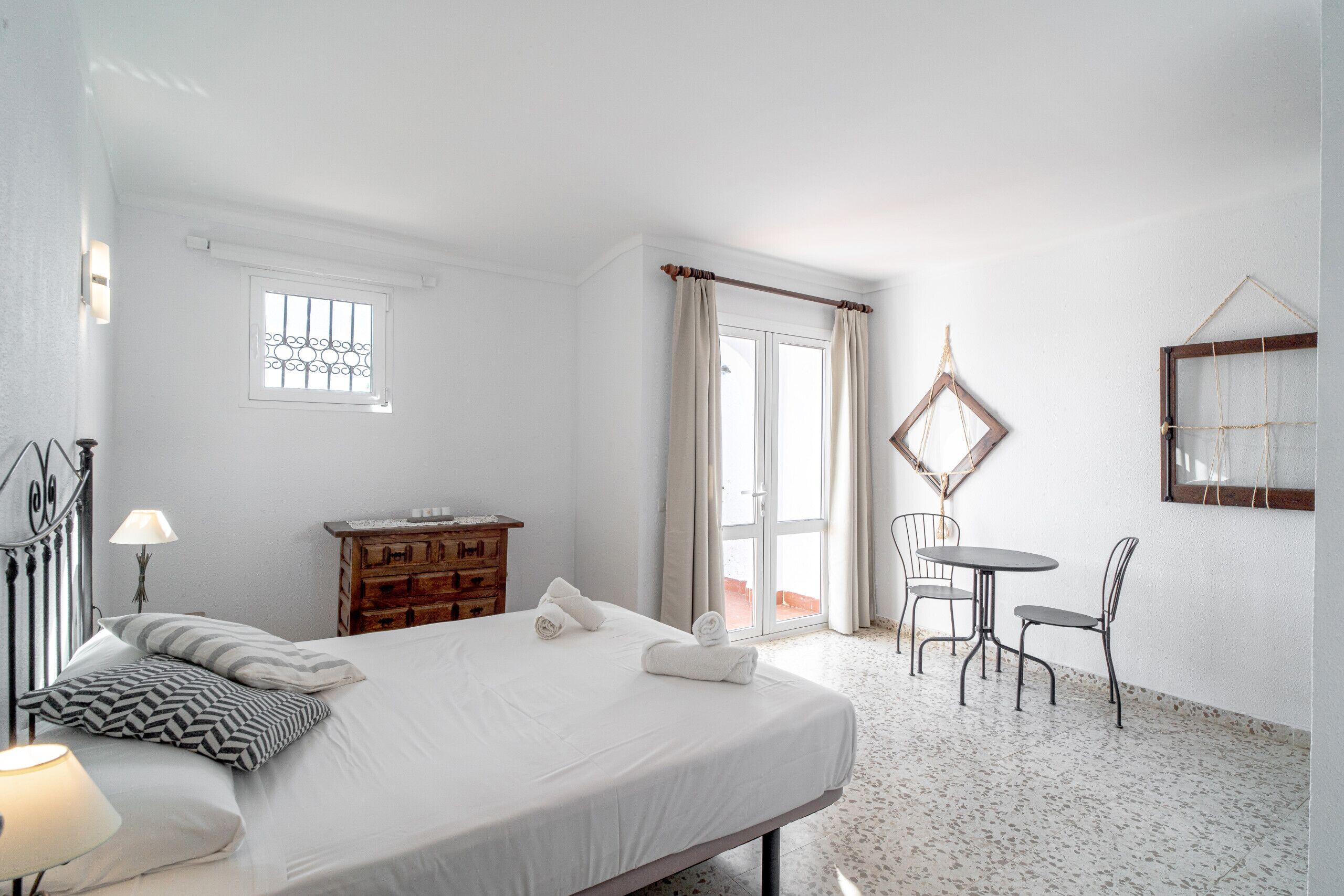 A bedroom with a bed, a wooden dresser, a table with chairs, a window, and a balcony door.
