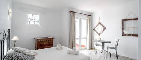 A bedroom with a bed, a wooden dresser, a table with chairs, a window, and a balcony door.