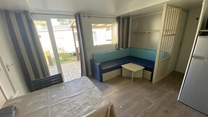 A compact room with a kitchenette, a seating area with a bench, a small table, and a sliding glass door leading to an outdoor area. - Bungalow 'Mobil-home Narbonne Plage' with Shared Pool, Private Terrace and Air Conditioning (Narbonne)