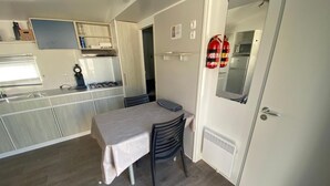 A compact kitchen with a stove, sink, and cabinets, a dining area with a table and chairs, and a door with a fire extinguisher. - Bungalow 'Mobil-home Narbonne Plage' with Shared Pool, Private Terrace and Air Conditioning (Narbonne)