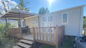 A mobile home with a wooden deck, a covered patio, and a car parked nearby. - Bungalow 'Mobil-home Narbonne Plage' with Shared Pool, Private Terrace and Air Conditioning (Narbonne)