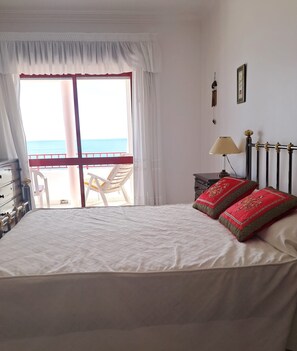 Ein Schlafzimmer mit einem Bett, einem Nachttisch, einer Lampe, einem Fenster mit Vorhängen und Blick aufs Meer.