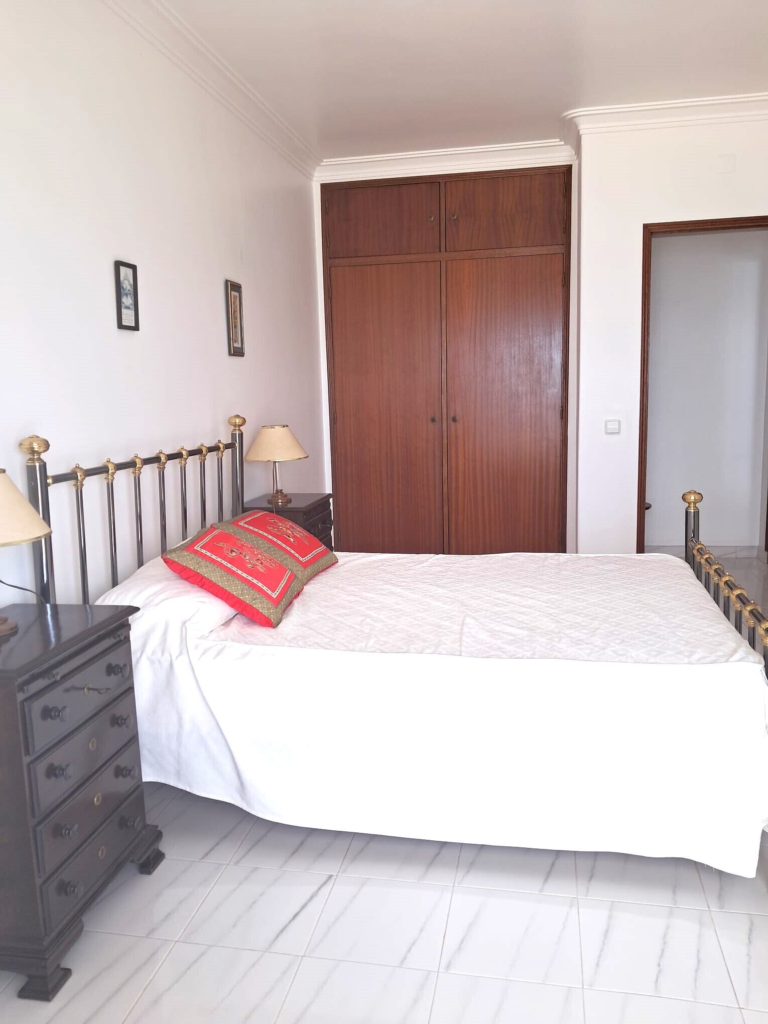 A bedroom with a bed, dresser, nightstand, lamp, and a large wooden wardrobe.