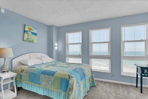 A bedroom with a bed, a nightstand, a lamp, a painting, and large windows. - 3 Bedroom Point Condo Unit (North Topsail Beach)