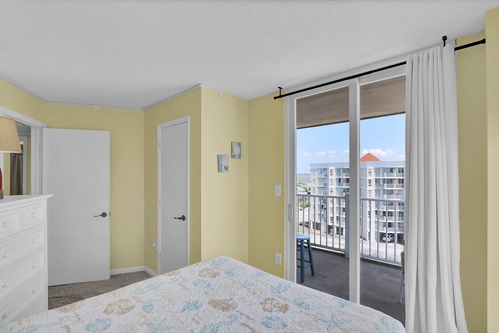 A bedroom with a bed, a dresser, a door, and a balcony with a view of buildings.