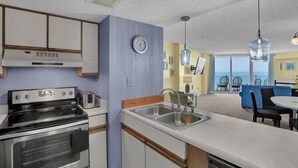 A compact kitchen with a stove, toaster, and sink, a clock, and a view of the balcony with chairs. - 3 Bedroom Point Condo Unit (North Topsail Beach)