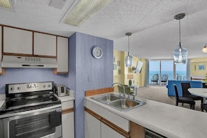 A compact kitchen with a stove, toaster, and sink, a clock, and a view of the balcony with chairs. - 3 Bedroom Point Condo Unit (North Topsail Beach)