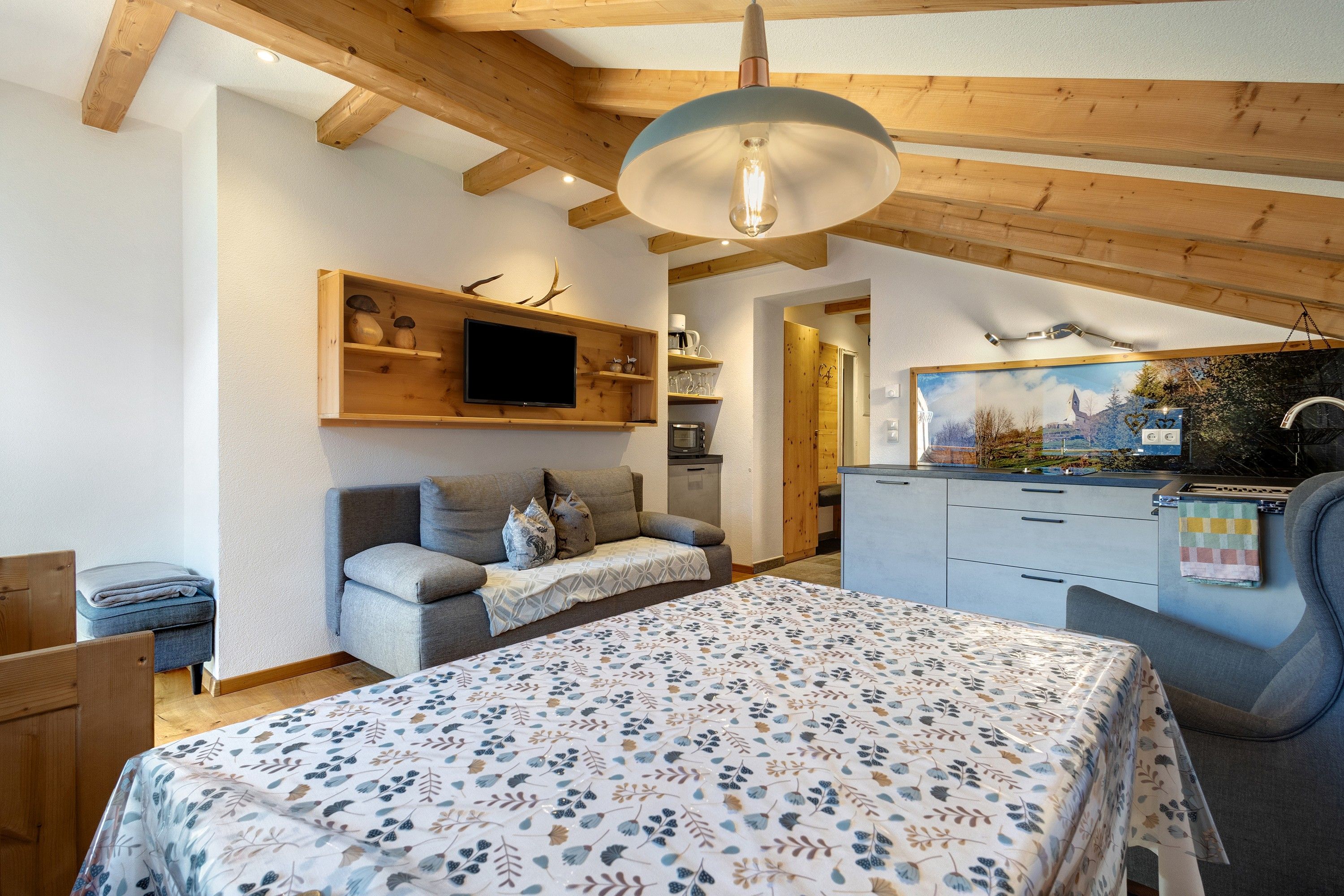 Una camera accogliente con un letto, un divano, una TV montata a parete e una cucina visibile attraverso una porta aperta.