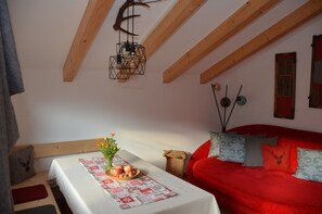 Ein gemütliches Zimmer mit einem roten Sofa, einem Holztisch mit Obstschale und Blumen, einer Pendelleuchte und einer Holzzieldecke.