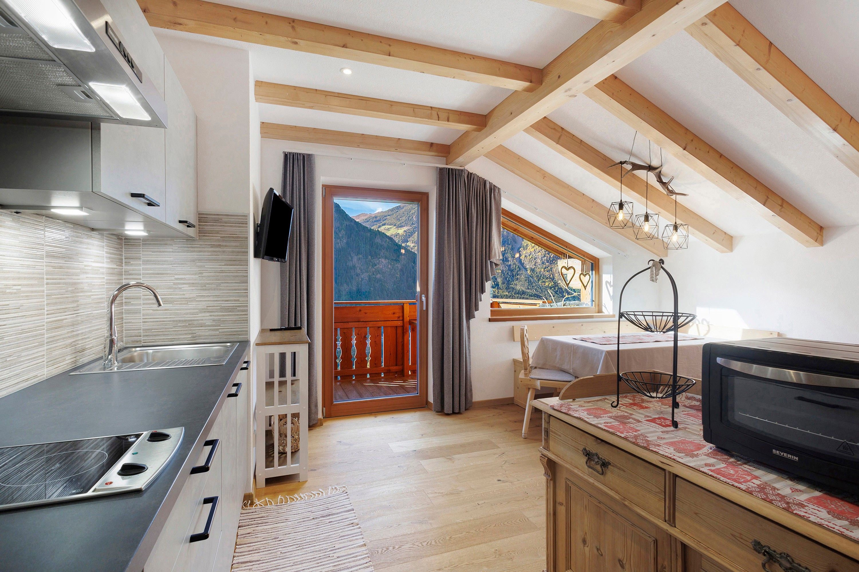 A modern kitchen with a built-in oven, a dining area with a table and chairs, and a balcony with a view of the mountains.
