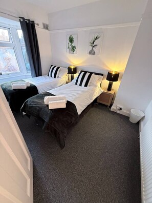 Ein Hotelzimmer mit zwei Betten, einem Fenster mit Vorhängen, einem Heizkörper und einer an der Wand befestigten Pflanze.