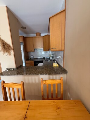 A compact kitchen with wooden cabinets, a granite countertop, and a dining area with a wooden table and chairs. - Apartamento Torreblanca III Ref 063 (Torreblanca)