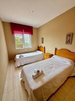 A bedroom with two beds, a window with a red blind, a wooden nightstand, and a small bedside table. - Apartamento Torreblanca III Ref 063 (Torreblanca)