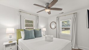 Ein Schlafzimmer mit Bett, Nachttischen, Deckenventilator und Fenstern mit Vorhängen.