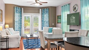 A hotel room with a ceiling fan, a dining table with chairs, a sofa, a TV, and a window with curtains. - On the Links-9th Hole- L egends Stay (Myrtle Beach)