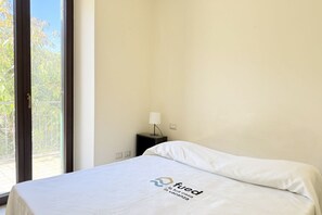 A hotel room with a bed, a nightstand, a lamp, and a view of greenery through the window.