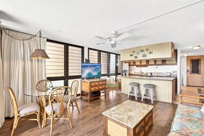 Ein Wohnzimmer mit Fernseher, Holzmöbeln, einem Deckenventilator und einer Küchenzeile mit Barhocker.