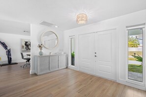 Ein modernes Interieur mit Holzboden, weißen Wänden, einem runden Spiegel, einer weißen Konsole und einer zur Außenseite führenden Tür.