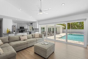 A modern living room with a sectional sofa, ottoman, and a view of a swimming pool through large sliding glass doors. - Morningside Retreat Home by RVA (Longboat Key)