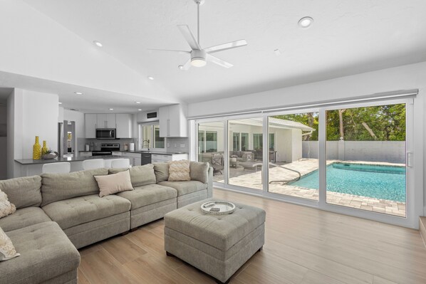 Ein modernes Wohnzimmer mit einem Ecksofa, einem Hocker und Blick auf einen Swimmingpool durch große Schiebetüren.