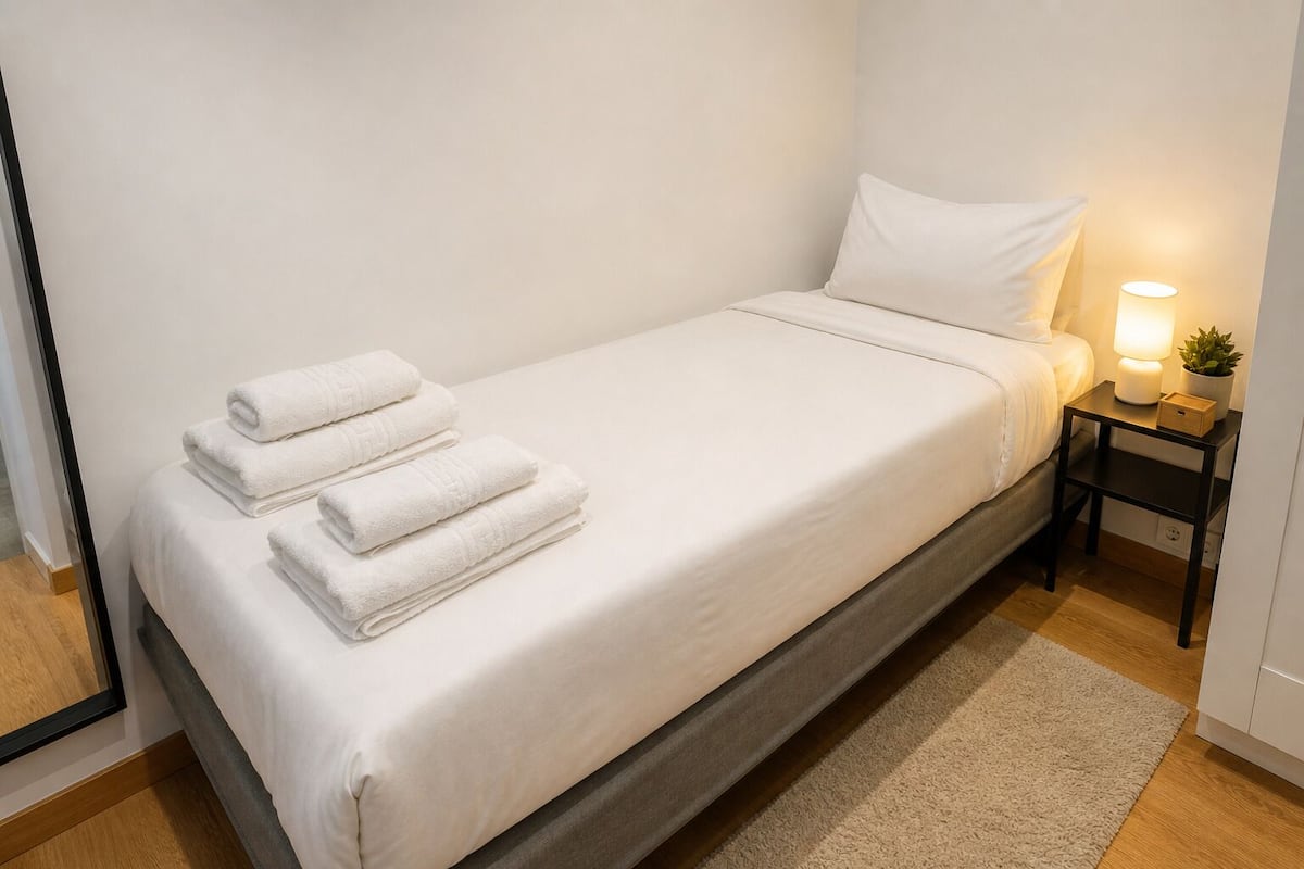 Een eenpersoonsbed met witte lakens, een bedlampje, een klein plantje en een spiegel aan de muur.