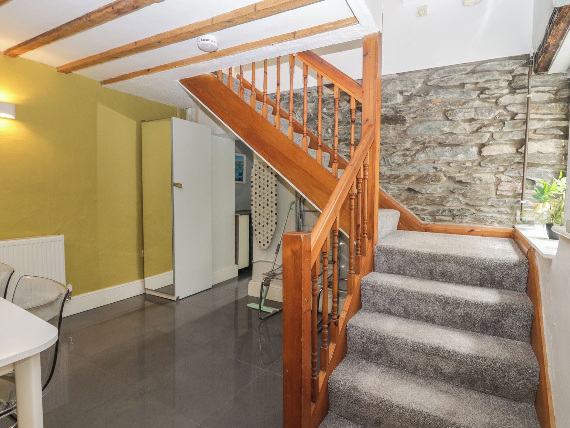 Eine Treppe mit Holzwägeln führt zu einer Zwischenebene, eine Steinwand und ein Küchenbereich mit einem Kühlschrank.