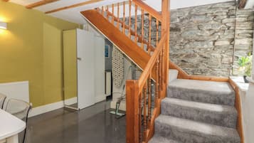 Eine Treppe mit Holzwägeln führt zu einer Zwischenebene, eine Steinwand und ein Küchenbereich mit einem Kühlschrank.