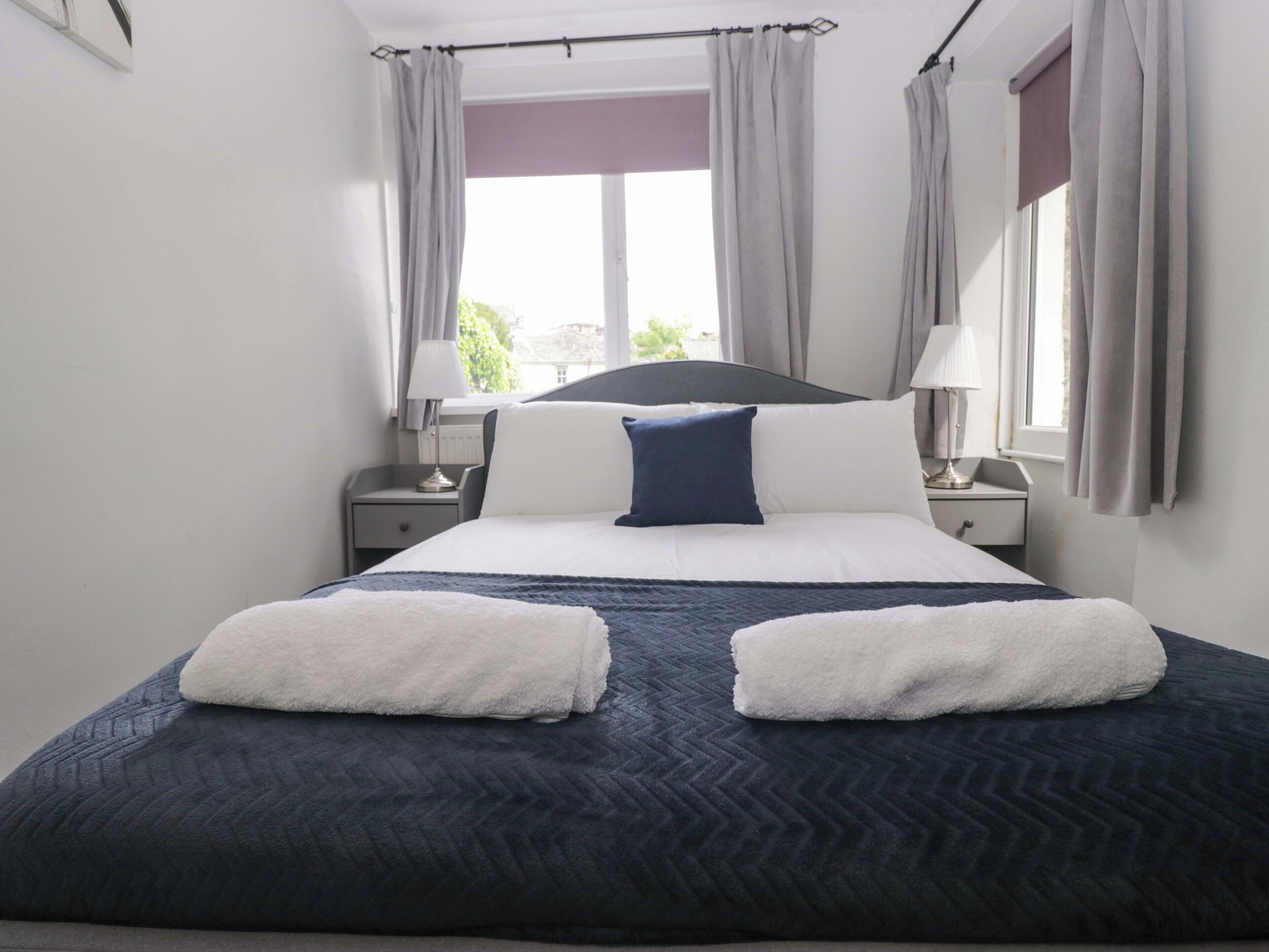 A neatly made bed with a dark blue quilt, white pillows, and two white towels. There are two bedside tables with lamps, a window with curtains, and a view of greenery outside.
