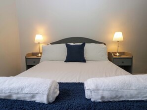 A neatly made bed with white and blue bedding, two bedside lamps, and two folded towels on the floor. - Courtyard Cottage (Grange-over-Sands)