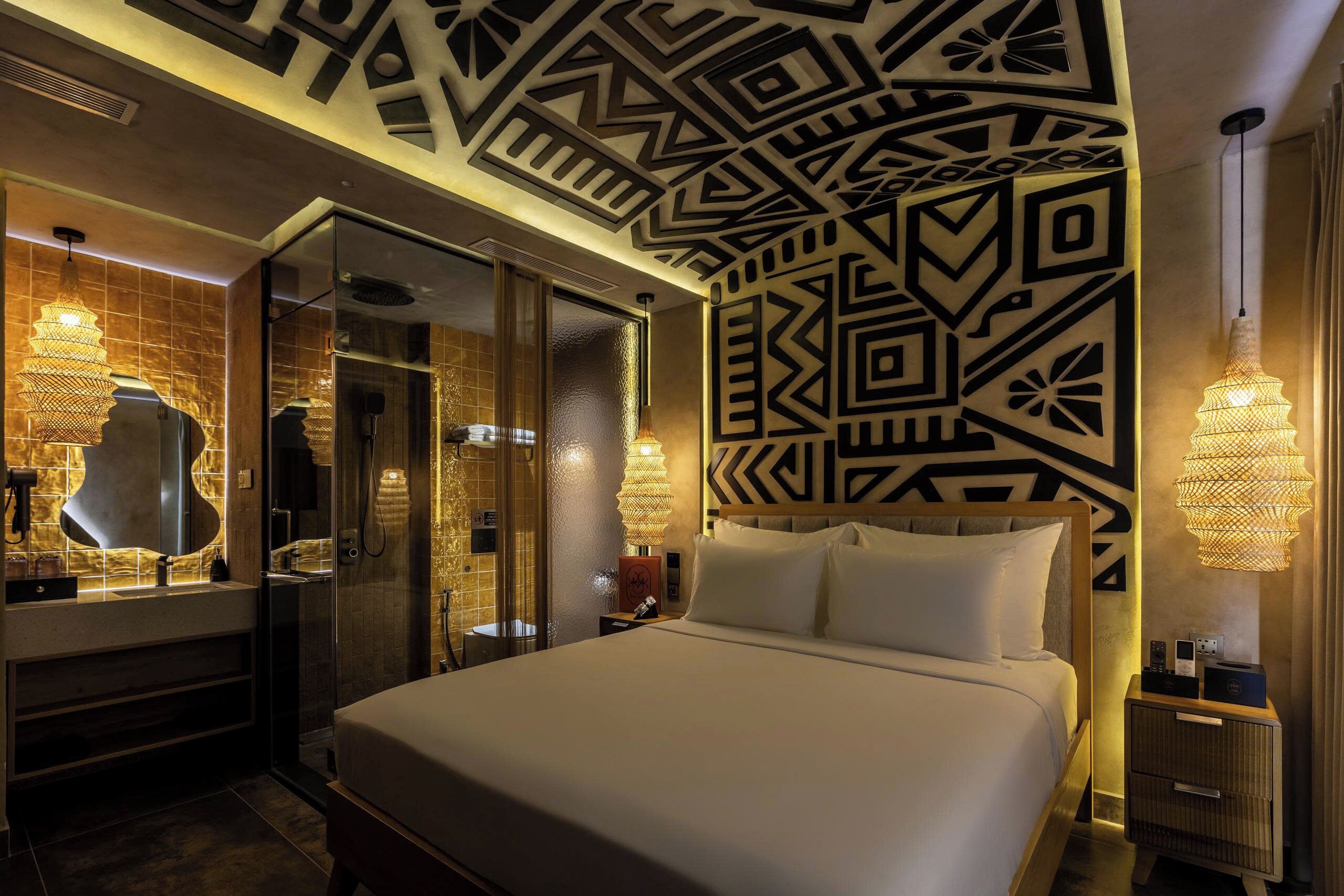 A hotel room with a bed, a bathroom with a shower, a vanity with a mirror, and a wall with geometric patterns.