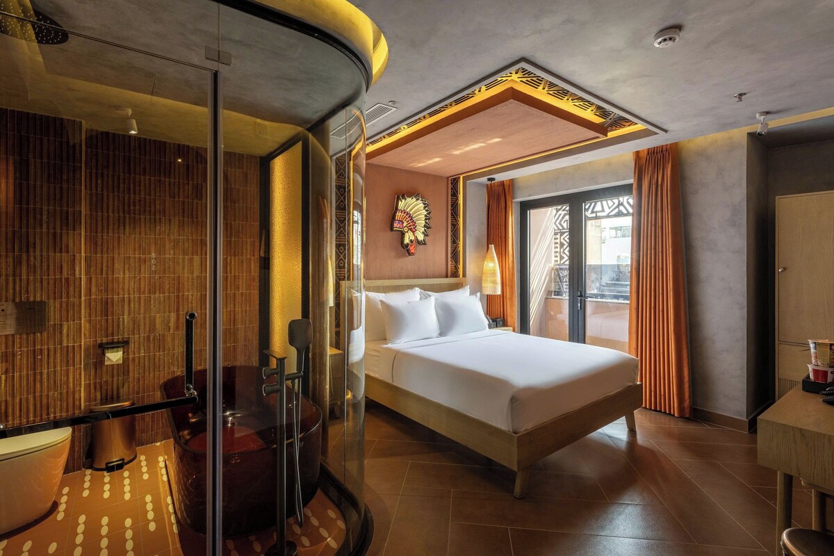 Habitación de hotel con una cama grande, cabina de ducha de vidrio, cabecero de madera y vistas al exterior.