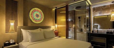 A modern hotel room with a large bed, a wall-mounted decorative plate, a glass shower enclosure, and a vanity with a mirror.