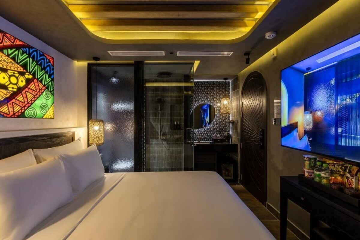 Habitación de hotel con cama, televisor, una obra de arte en la pared y baño visible a través de una puerta de cristal.