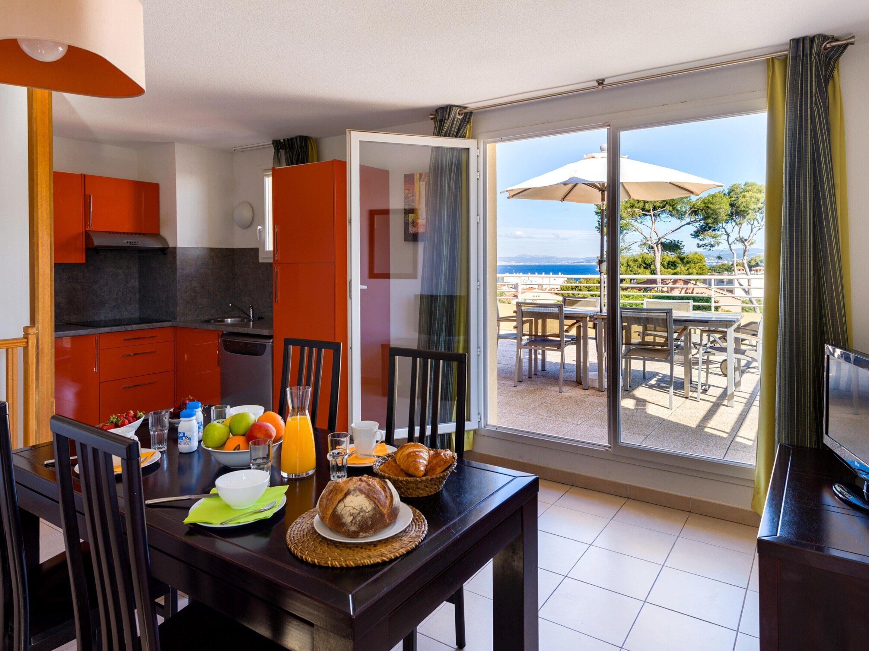 A dining area with a table set for breakfast, a kitchen with orange cabinets, and a balcony with outdoor seating.