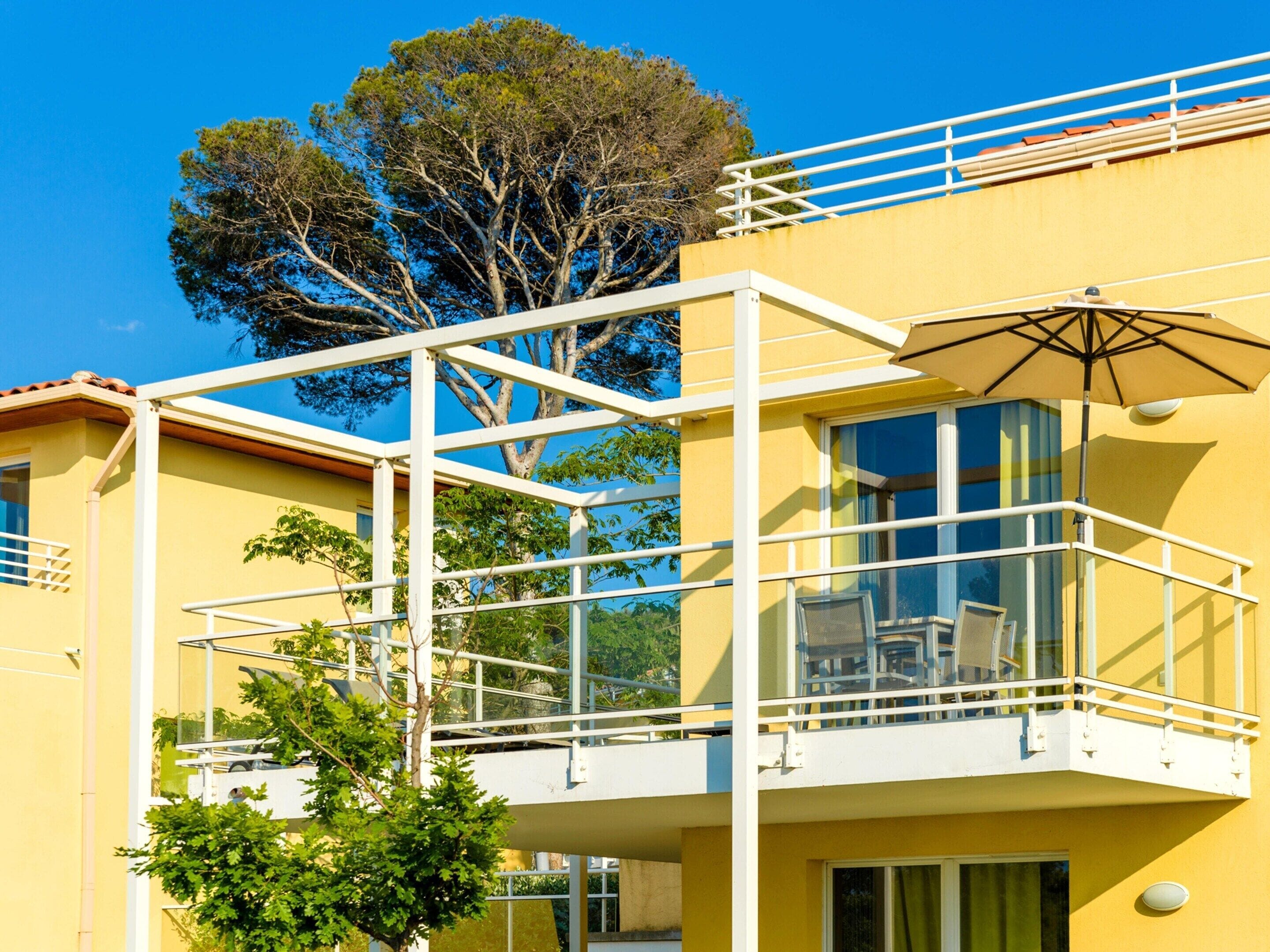 A yellow building with a balcony, glass railings, and a tree in the background.