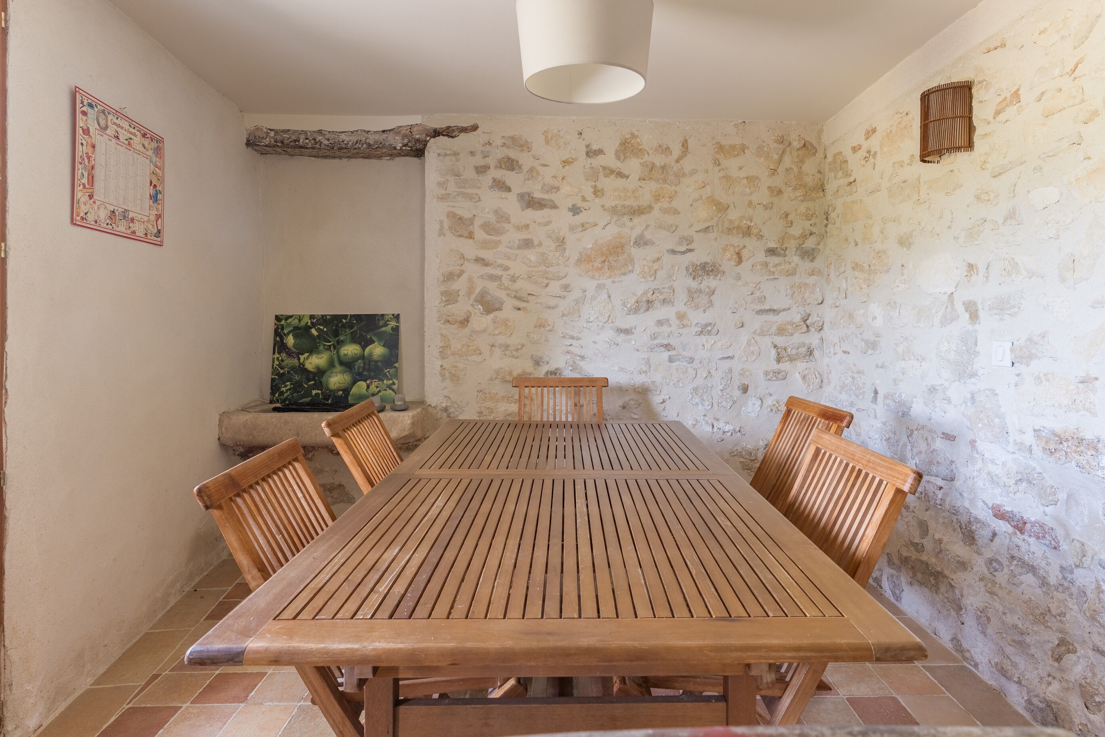 Un coin repas avec une table et des chaises en bois, un mur en pierre, un luminaire suspendu et un tableau encadré au mur.