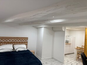 Ein Schlafzimmer mit einem Bett, einem Holzkopfteil, einem kleinen Hocker und einer Küchenzeile mit Kühlschrank und Wasserkocher.
