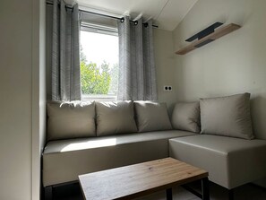 A modern living room with a sofa, a wooden coffee table, and a window with curtains. - Mobil-home, pets allowed - 6 guests (Saint-Martin-Lars-en-Sainte-Hermine)