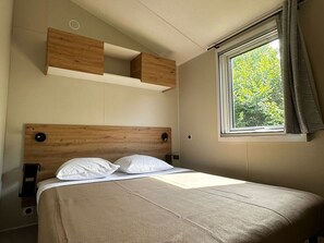 A bedroom with a wooden headboard, a bed with white bedding, a window with curtains, and a built-in shelf above the bed. - Mobil-home, pets allowed - 6 guests (Saint-Martin-Lars-en-Sainte-Hermine)