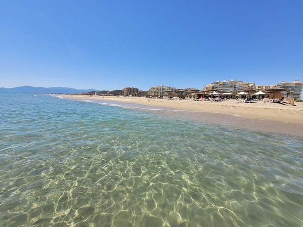 Ein Strand mit klarem Wasser, sandigem Ufer und Gebäuden im Hintergrund.