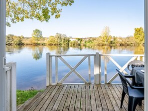 Una terraza de madera con una parrilla, con vista a un lago tranquilo con árboles y casas al fondo.
