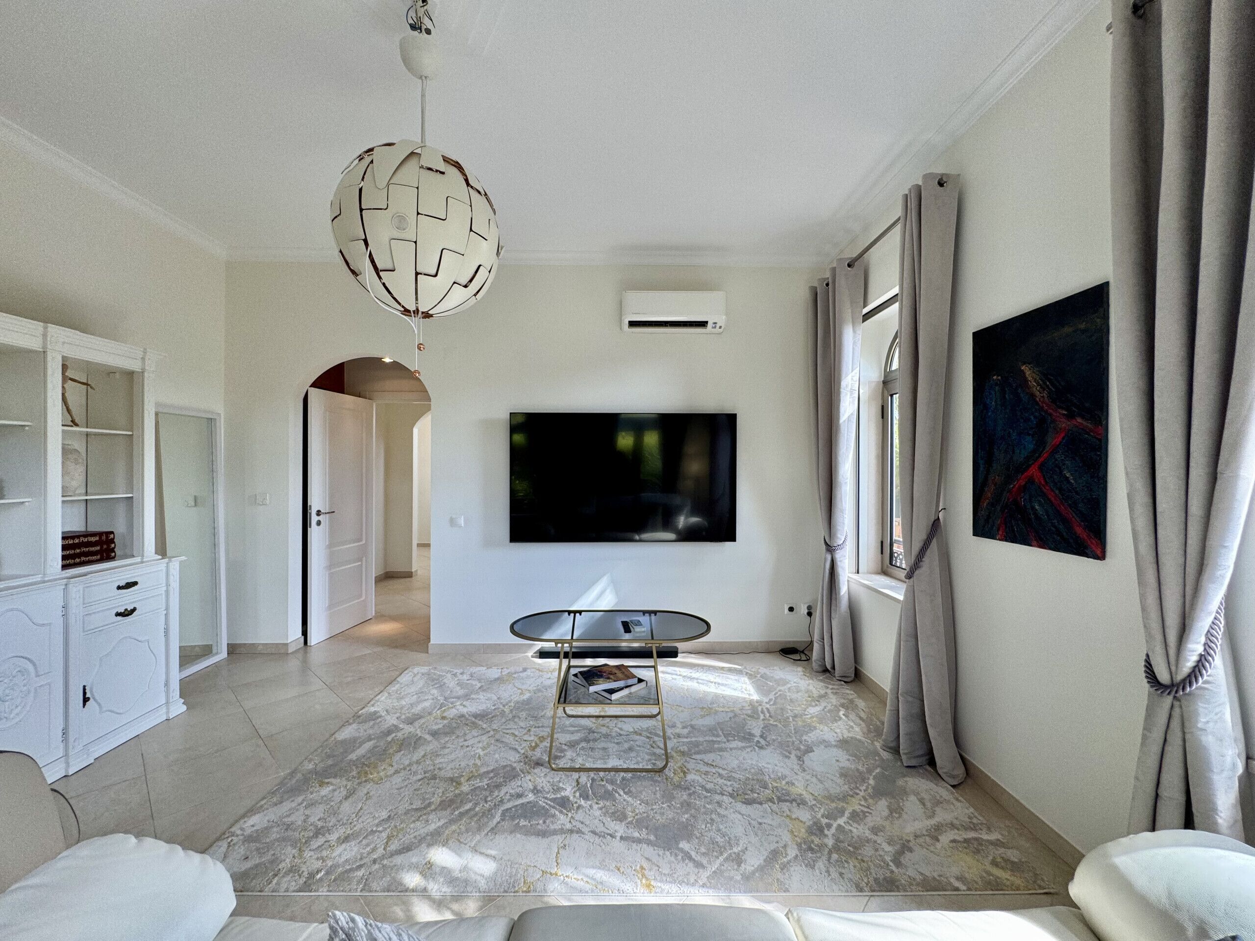 Een moderne woonkamer met een flatscreen televisie, een vloerkleed met marmerprint, een wit tv-meubel en een groot raam met gordijnen.