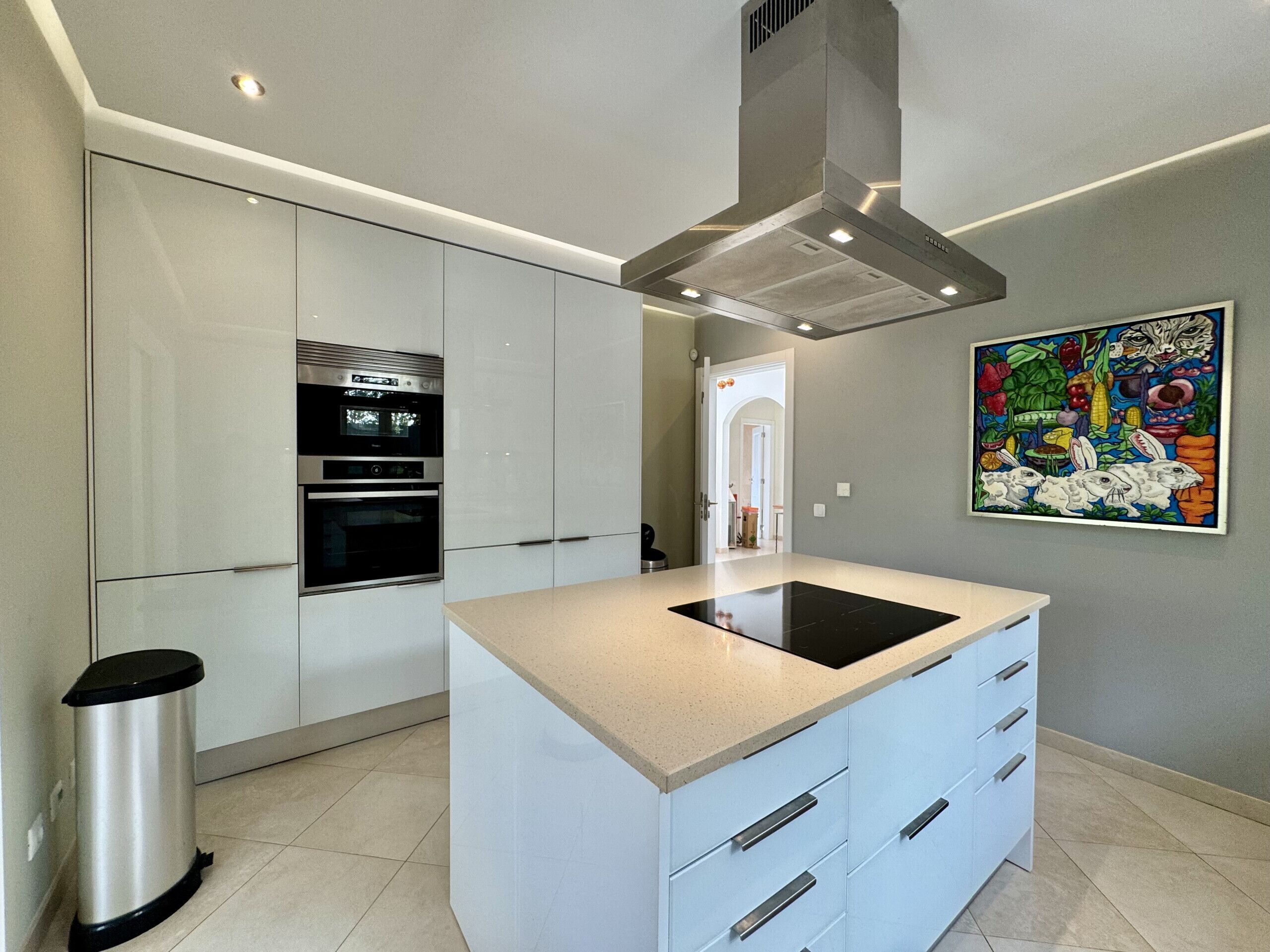 Une cuisine moderne avec des meubles blancs, une hotte en acier inoxydable et un îlot central doté d’une plaque de cuisson à induction noire.