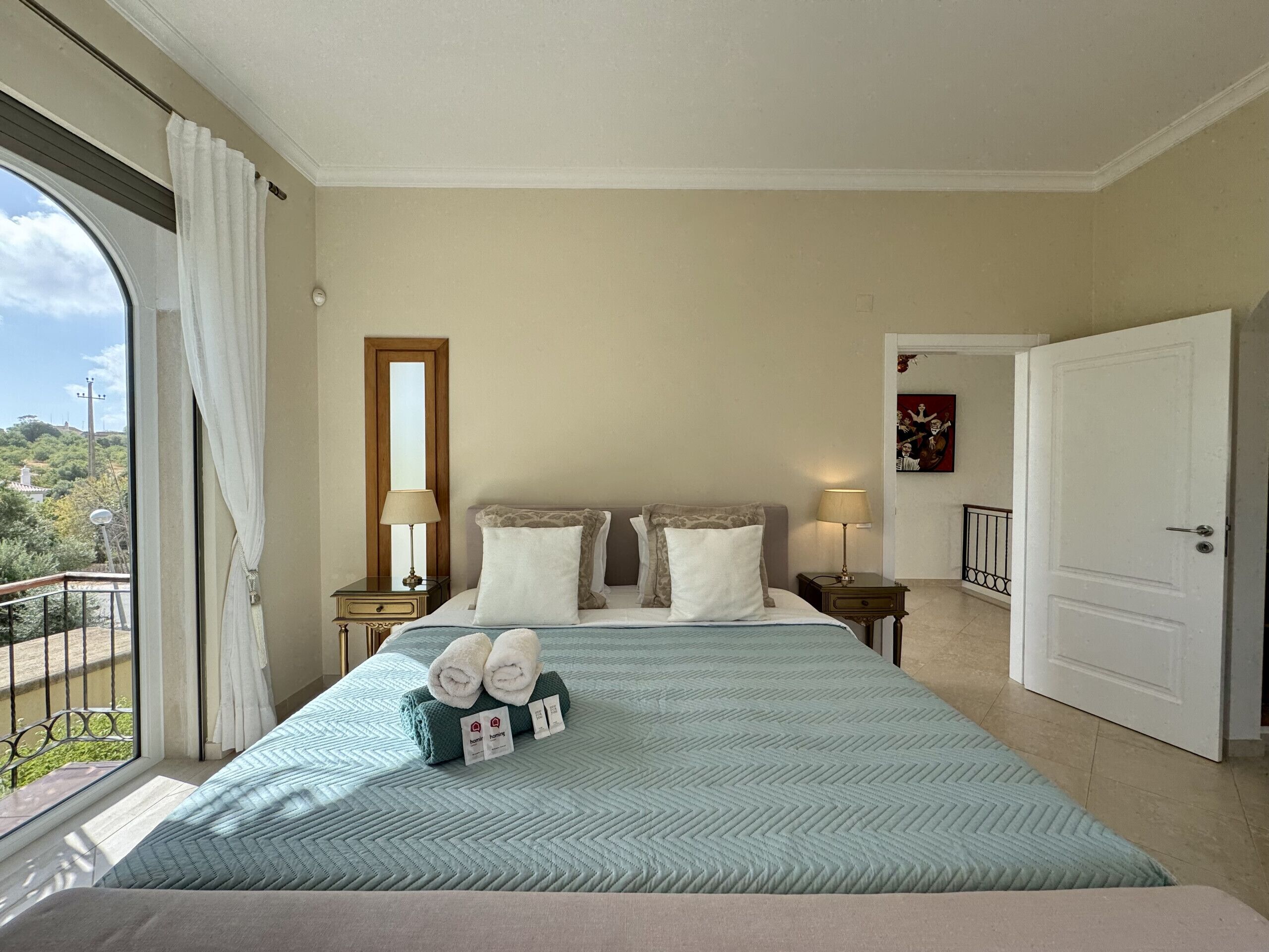A hotel room with a bed, two bedside tables, a window with curtains, a door, and a view of the outside.