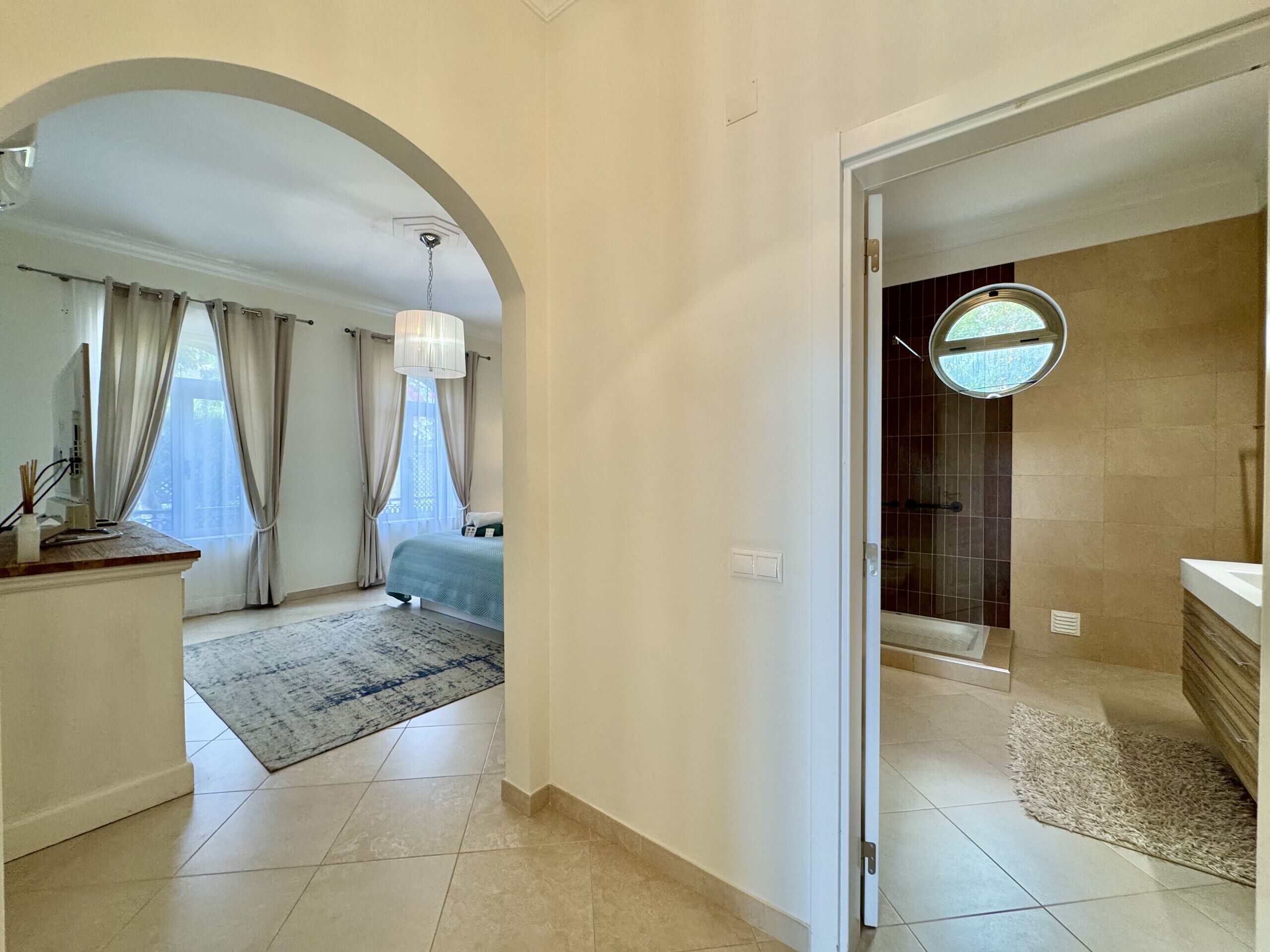 A hotel room with a bed, a desk, a window with curtains, and a bathroom with a shower and sink.